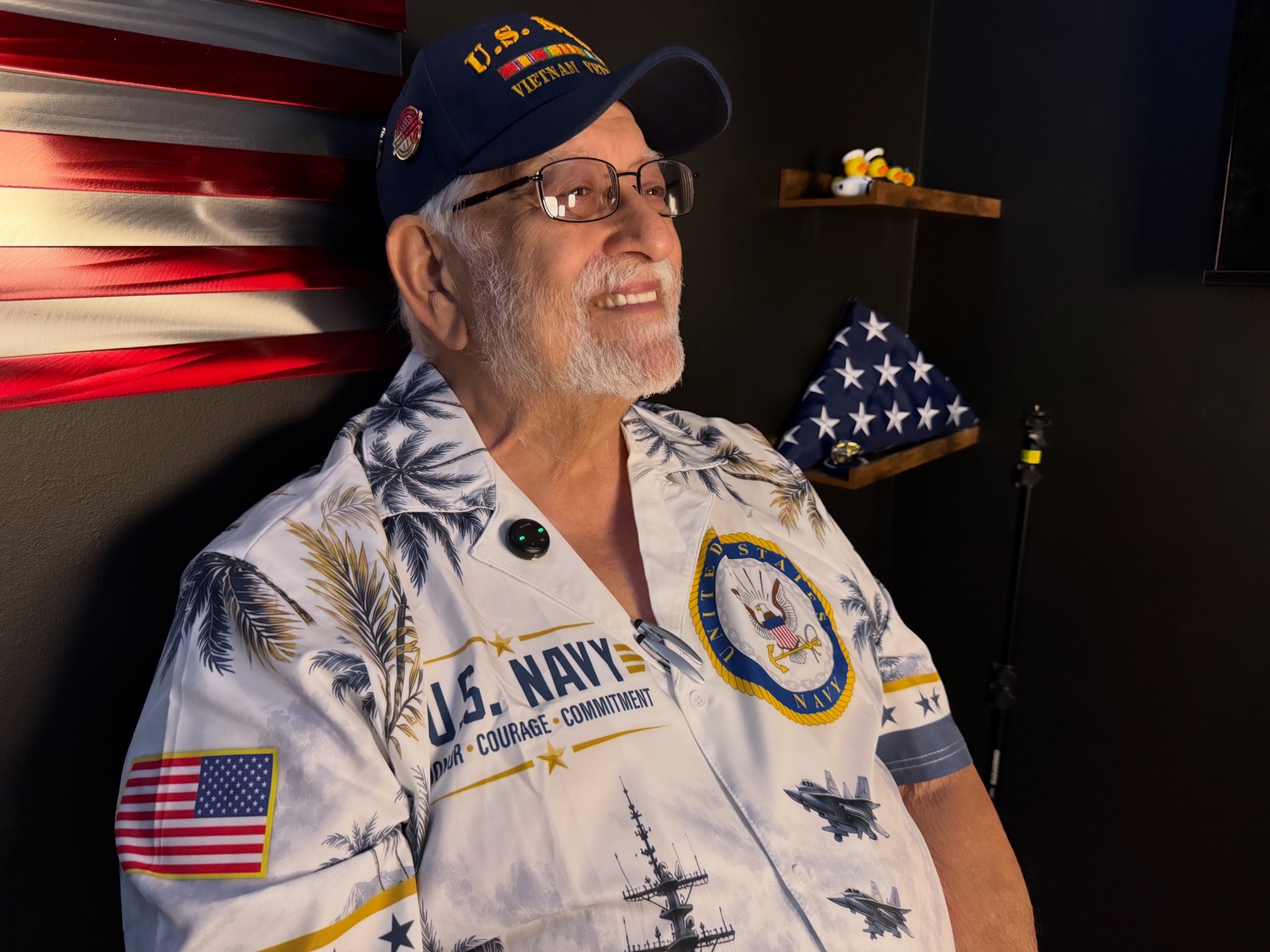 Mike Martin smiling in his U.S. Navy Vietnam veteran cap beside a mounted American flag